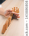 A hand is positioned at the end of a diagonal line of leaning wooden domino blocks, with wooden peg figures beyond it, creating a vertical concept image of a barrier against a potential 143886808