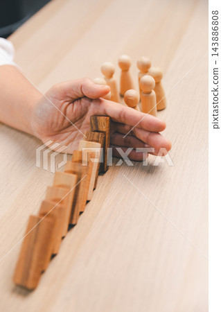 A hand is positioned at the end of a diagonal line of leaning wooden domino blocks, with wooden peg figures beyond it, creating a vertical concept image of a barrier against a potential A hand is positioned at the end of a diagonal line of leaning wooden domino blocks, with wooden peg figures beyond it, creating a vertical concept image of a barrier against a potential 143886808