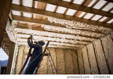 Professional worker applies spray insulation to upper structure of timber home Professional worker applies spray insulation to upper structure of timber home 143888196