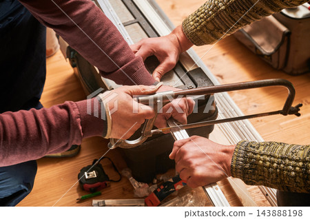 Two men cutting metal profile using hand hacksaw, holding rail steady during precise trimming Two men cutting metal profile using hand hacksaw, holding rail steady during precise trimming 143888198