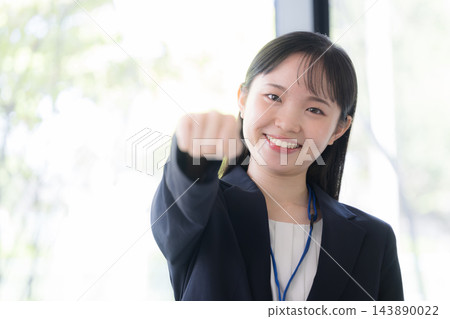 一位二十出頭的日本年輕女性，身穿西裝，戴著員工證，直視鏡頭，擺出勝利的姿勢並揮拳。 143890022