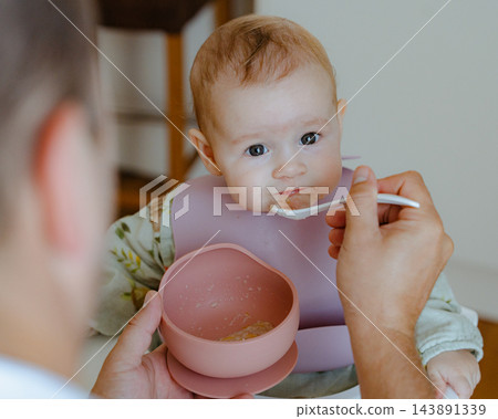 Cute baby girl looking curiously into the camera while being fed by her father, highlighting early childhood nutrition, tender parent care, and love Cute baby girl looking curiously into the camera while being fed by her father, highlighting early childhood nutrition, tender parent care, and love 143891339