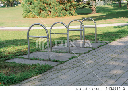 Outdoor stainless steel bike stands located in a quiet green park along a paved footpath under bright sunlight, representing cycling infrastructure Outdoor stainless steel bike stands located in a quiet green park along a paved footpath under bright sunlight, representing cycling infrastructure 143891340