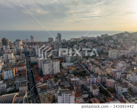 Durres Albania appears in broad coastal panorama at sunset, with dense urban buildings beside Adriatic Sea. Tall towers, wooded hillside and distant ships distinguish this seaside city view. 143891442