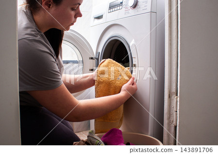 Woman loads pile of dirty clothes into modern washing machine, using home appliances to simplify laundry. Automated washing makes household routines more convenient and efficient Woman loads pile of dirty clothes into modern washing machine, using home appliances to simplify laundry. Automated washing makes household routines more convenient and efficient 143891706