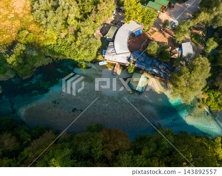 Blue Eye Albania natural spring winds through dense woodland, its brilliant blue pools bordering pebbled riverbed. Overhead perspective includes visitor buildings beside this striking scenic waterway. Blue Eye Albania natural spring winds through dense woodland, its brilliant blue pools bordering pebbled riverbed. Overhead perspective includes visitor buildings beside this striking scenic waterway. 143892557