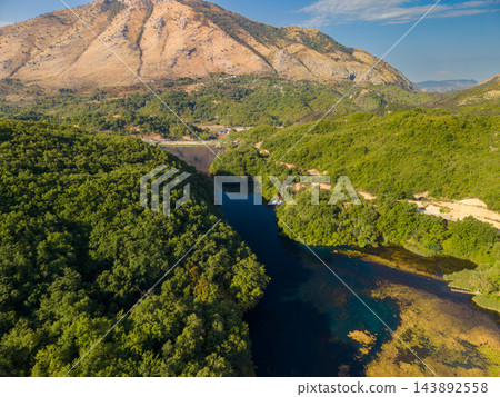 Blue Eye Albania spring sits within broad green valley beneath rocky slopes. Forested banks frame deep blue water, creating tranquil natural landmark in Albanias mountain landscape. 143892558