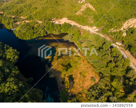 Blue Eye Albania spring lies amid dense green woodland, its deep blue water edged by golden vegetation. High aerial perspective shows surrounding hills, curving road and small riverside structure. Blue Eye Albania spring lies amid dense green woodland, its deep blue water edged by golden vegetation. High aerial perspective shows surrounding hills, curving road and small riverside structure. 143892559