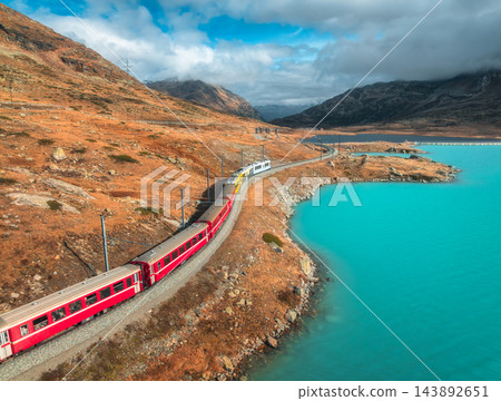 Aerial view of red train near alpine mountain lake and cloudy sky Aerial view of red train near alpine mountain lake and cloudy sky 143892651