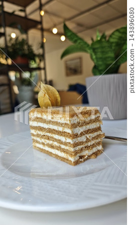 Layered vanilla cream cake slice with golden sponge, textured topping, and dried fruit garnish on a white ceramic plate in a softly lit restaurant interior 143896080