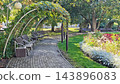 Curving brick walkway with benches beneath leafy trees, arched garden lights, green lawn, and colorful flower beds in a tranquil landscaped public park setting outdoors 143896083