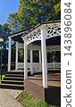 Ornate white wooden gazebo with decorative cutout railings, dark roof, steps, and open sides among mature green trees beside a paved walkway in a landscaped park setting 143896084