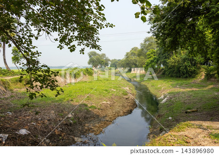 Narrow rural water canal flowing through green fields and trees. Scenic countryside stream landscape in a calm outdoor environment. 143896980