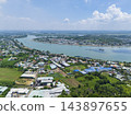 Aerial drone view of a Vietnamese riverside town with cargo barges on a winding river, flanked by residential areas and lush greenery under a blue sky. 143897655