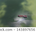 Aerial drone top-down view of a red barge navigating green river water, with cloud shadows and sunlight creating a striking contrast across the surface. 143897656