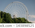 A giant white Ferris wheel stands beyond the green trees. 143898525