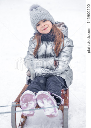 Little girl smiling on a wooden sled in snow wearing a silver winter coat and knit hat Little girl smiling on a wooden sled in snow wearing a silver winter coat and knit hat 143900200