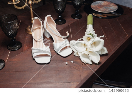 White strappy bridal sandals with rhinestone straps arranged beside calla lily bouquet pearl earrings and gold ring on wooden table with dark glass goblets and decorative accents White strappy bridal sandals with rhinestone straps arranged beside calla lily bouquet pearl earrings and gold ring on wooden table with dark glass goblets and decorative accents 143900653