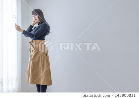 一位年輕女子正在窗邊對著手鏡整理頭髮。 143900831