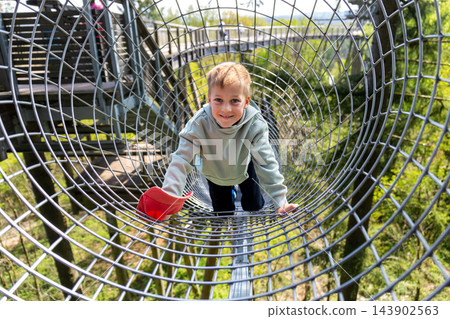 Cute little funny caucasian blond kid boy enjoy having fun walk metal net tunnel playground in treetop adventure amusement park Saarland. Children outdoors nature extreme sport activities concept 143902563