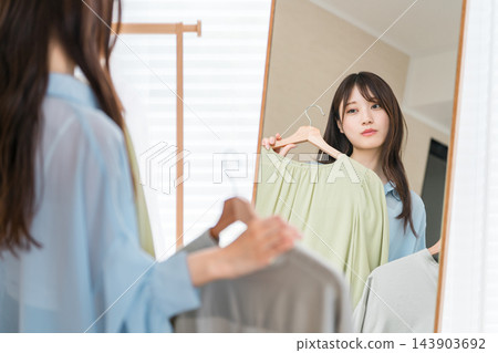 一位年輕女子對著鏡子比較不同的衣服，難以決定要穿什麼衣服。 143903692