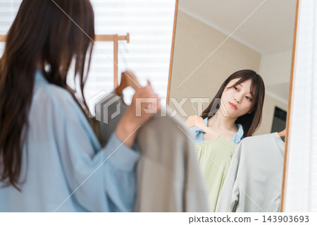一位年輕女子對著鏡子比較不同的衣服，難以決定要穿什麼衣服。 143903693