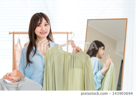 一位微笑的女士在家中拿著掛在衣架上的兩件衣服。 143903694