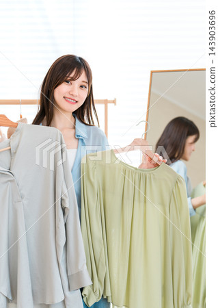 一位年輕女子麵帶微笑，手裡拿著兩套衣服，站在她家的鏡子旁。 143903696