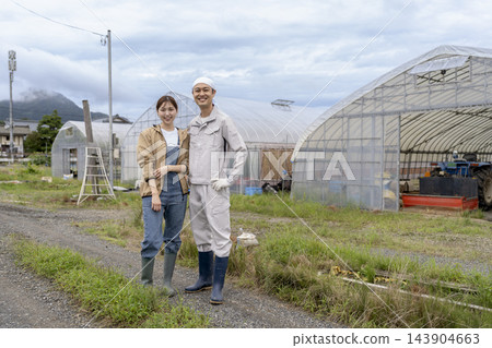 一位面帶微笑的農夫和他的妻子站在溫室前。 143904663
