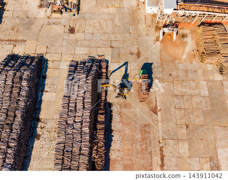 Aerial view of a forklift moving timber stacks across a paved lumber yard during daytime Aerial view of a forklift moving timber stacks across a paved lumber yard during daytime 143911042