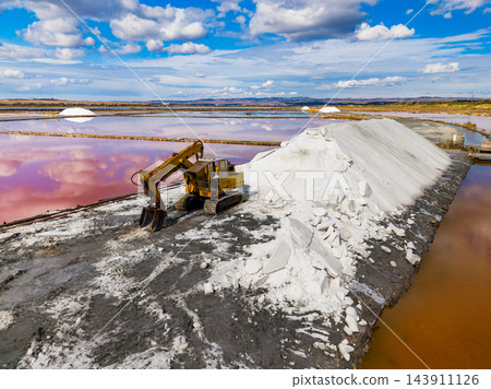 Excavator gathers salt beside pink evaporation ponds at a salt extraction site under a clouded sky Excavator gathers salt beside pink evaporation ponds at a salt extraction site under a clouded sky 143911126
