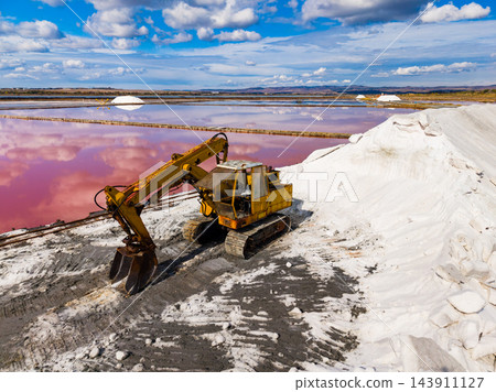 Excavator moves salt beside a pink evaporation pond at a salt extraction site under a cloudy daytime sky Excavator moves salt beside a pink evaporation pond at a salt extraction site under a cloudy daytime sky 143911127