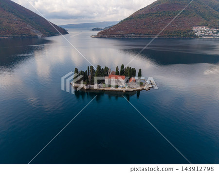 Small island church rests offshore near Perast, Montenegro, surrounded by dark blue water. Aerial landscape includes red roofed stone buildings, cypress trees, distant hills and coastal homes. Small island church rests offshore near Perast, Montenegro, surrounded by dark blue water. Aerial landscape includes red roofed stone buildings, cypress trees, distant hills and coastal homes. 143912798