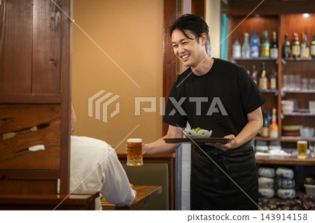 一位40多歲、微笑的男子在居酒屋(日本酒吧)為客人提供啤酒和食物。 一位40多歲、微笑的男子在居酒屋(日本酒吧)為客人提供啤酒和食物。 143914158