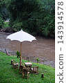 Wooden tables and chairs with white sun umbrellas are set up by the stream in front of the resort offering a scenic view in Thailand. Vertical photo 143914578