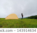 Smiling Asian woman standing orange tent camping on green expanse of grass on the mountain peak and see view greenery forest for lonely with peaceful calm in rain season 143914634