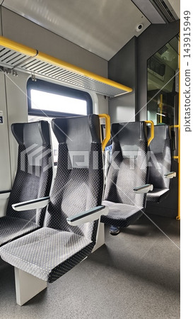 Empty modern public train seats with patterned upholstery, gray flooring, yellow handrails, bright window, and sleek interior fixtures inside a contemporary passenger rail carriage 143915949