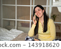 Smiling young woman speaking on a smartphone while sitting in a bright bedroom at home. Mobile voice call telecommunications and personal connection 143918620