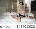 Smiling woman wrapped in a soft blanket while sitting on a bed in a bright bedroom. Home energy conservation personal warmth and cozy living 143918622