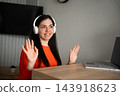 Smiling woman wearing headphones waves during a video meeting from home beside an open laptop. Remote collaboration online communication and home office work 143918623
