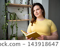 Young woman holding an open book beside indoor plants in a bright home interior. Digital wellbeing screen-free reading break and mindful relaxation 143918647