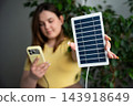 Woman holding portable solar panel and smartphone beside indoor greenery. Off grid power renewable energy and mobile charging 143918649
