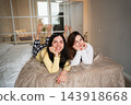 Two young women relaxing together on a bed in a bright modern home bedroom. At-home sleepover friendship social leisure and cozy weekend lifestyle 143918668