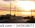 Empty elevated road crosses industrial district under glowing orange sunset, lined with tall streetlights and factories. Warm evening sky silhouettes warehouses, railings and urban infrastructure 143918673