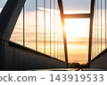 Close view of arched steel railway bridge in Magdeburg, Germany, silhouetted against glowing sunset sky. Vertical suspension rods and dark metal girders frame warm evening light above empty tracks 143919533