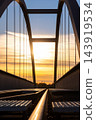 Close view of arched steel railway bridge in Magdeburg, Germany, silhouetted against glowing sunset sky. Vertical suspension rods and dark metal girders frame warm evening light above empty tracks 143919534
