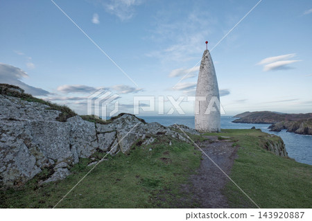 Tall white monument standing on a rocky cliff by the blue sea under a cloudy sky during the daytime. Tall white monument standing on a rocky cliff by the blue sea under a cloudy sky during the daytime. 143920887