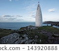 Tall white stone monument standing on a grassy cliff overlooking the blue ocean during the daytime. 143920888