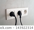 White power outlet panel mounted on a textured wall with two black plugs inserted, cables hanging and one empty socket visible. Conveys electricity, home or office interior, connection and everyday ut 143922314
