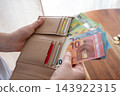Hands holding an open beige wallet filled with euro banknotes and credit cards, suggesting cash withdrawal, personal finances, budgeting, travel expenses and everyday spending in a warm home setting.  143922315